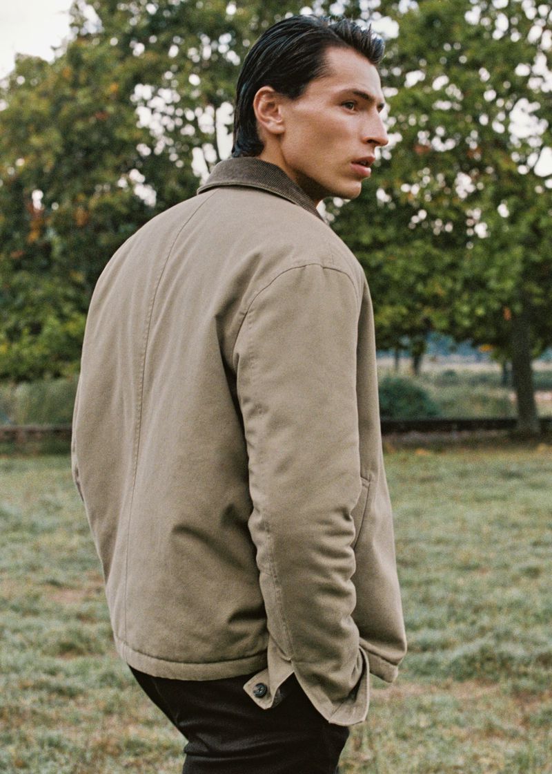 Man in tan jacket stands in a grassy field near trees, looking away from the camera. Neutral tones and natural light create a casual style.