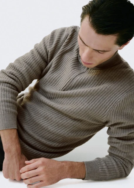 Man in ribbed knit sweater poses indoors against a white backdrop. He is angled down, gazing downward, with hands visible.