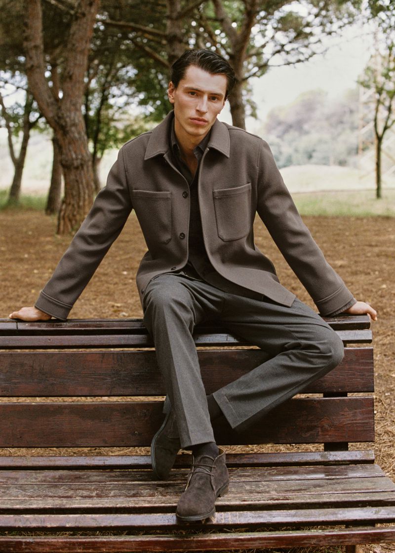 Male model poses on wooden bench in outdoor fashion shot. He is wearing a dark gray shirt, suit pants, matching jacket and suede boots.