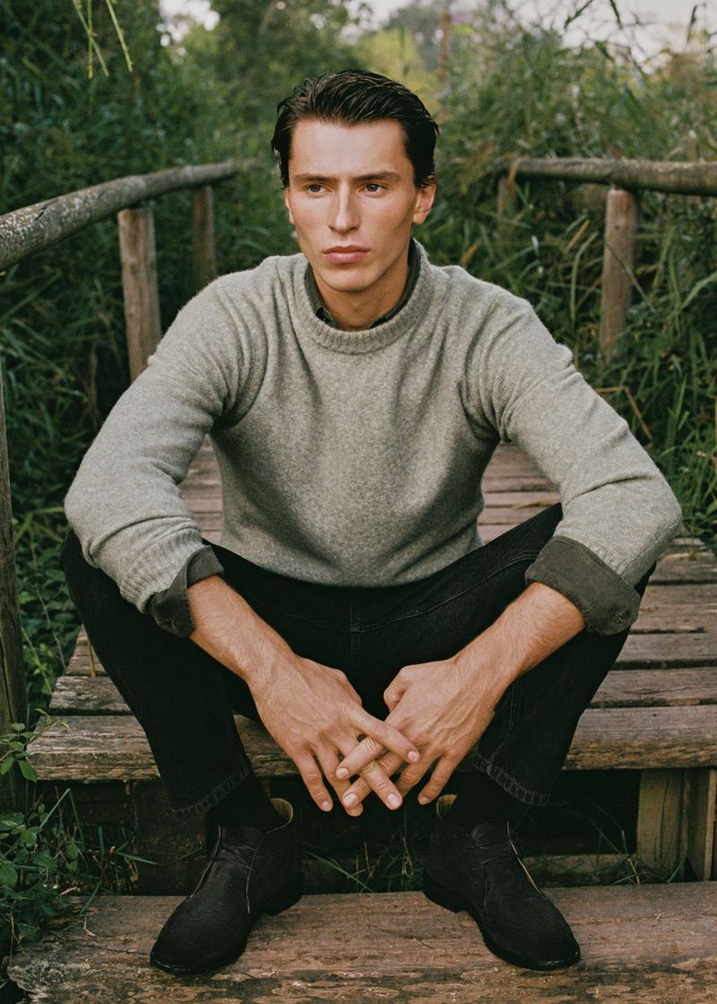 Portrait of a handsome adult male sitting on a wooden bridge in nature. He is wearing a grey sweater, dark denim jeans, and suede boots.
