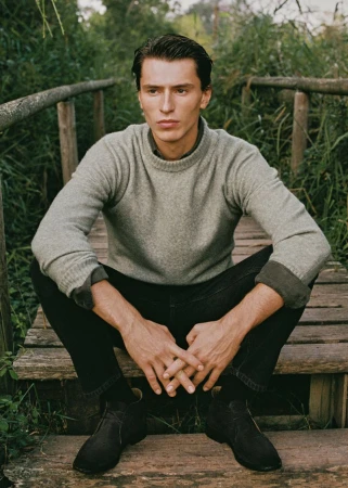 Portrait of a handsome adult male sitting on a wooden bridge in nature. He is wearing a grey sweater, dark denim jeans, and suede boots.
