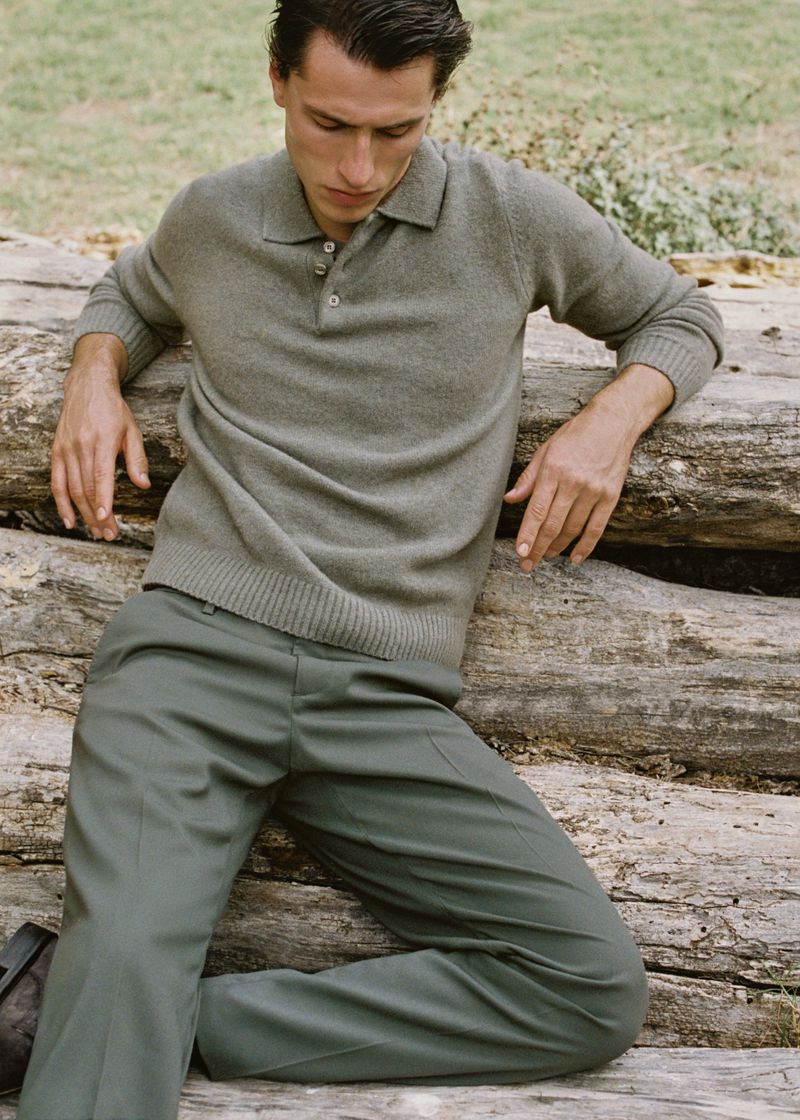 A male model in a polo and pants leans against logs in an outdoor setting, showcasing fashion and style with a natural backdrop.