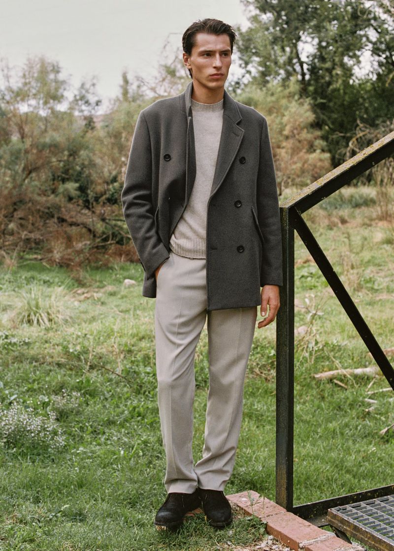 A stylish man in a gray jacket and beige pants stands outdoors on a sunny day, posing near a metal barrier surrounded by greenery.