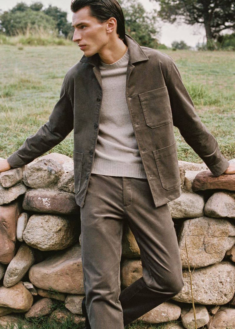 A stylish adult male poses confidently in a corduroy jacket and trousers outdoors, leaning against a rustic stone wall in a serene setting.