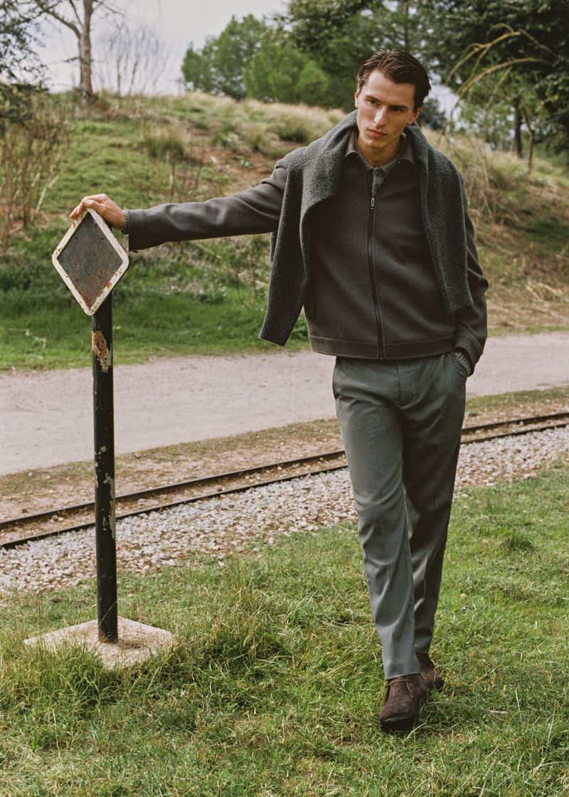 Male model poses outdoors in a gray jacket, trousers, and sweater, leaning against a sign near train tracks on a grassy hillside.