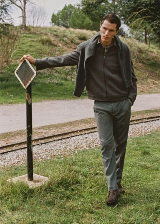 Male model poses outdoors in a gray jacket, trousers, and sweater, leaning against a sign near train tracks on a grassy hillside.