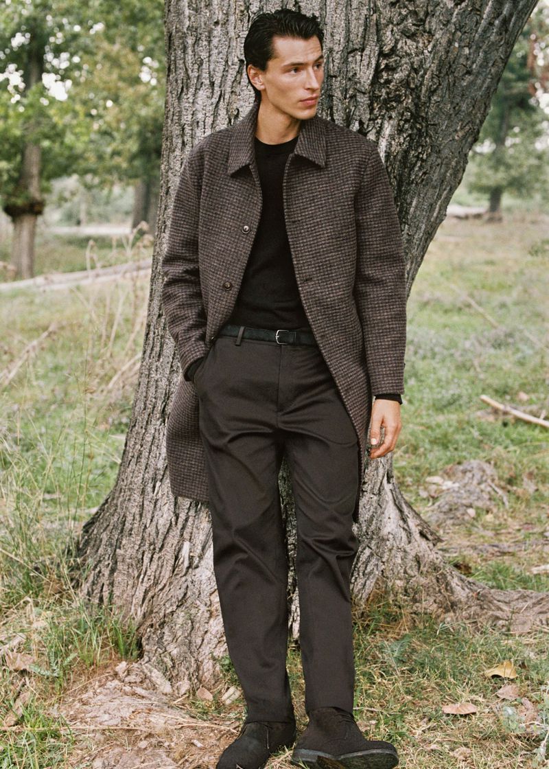 A young man in a coat stands in an outdoor setting near a tree. He is wearing casual attire with nature in the soft-focus background.