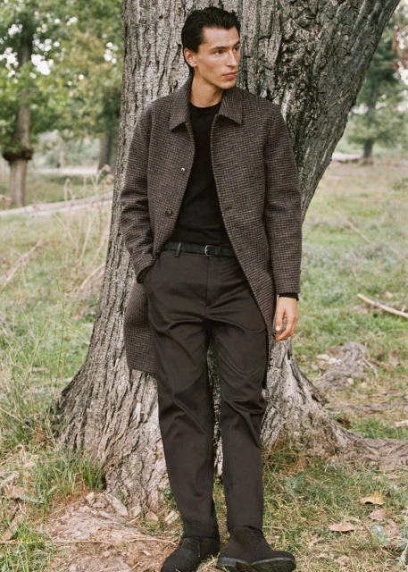A young man in a coat stands in an outdoor setting near a tree. He is wearing casual attire with nature in the soft-focus background.