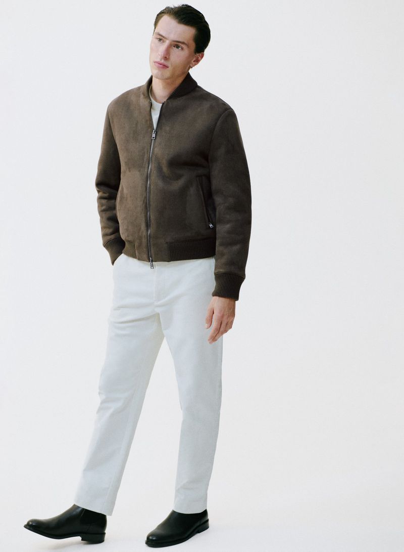 Full-length shot of a young adult man in a brown suede jacket, white pants, and black boots, standing against a plain white background.