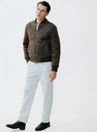 Full-length shot of a young adult man in a brown suede jacket, white pants, and black boots, standing against a plain white background.