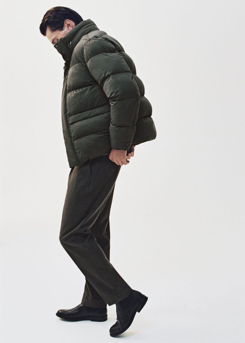 A man in a puffer jacket, trousers, and boots poses indoors against a white backdrop. He's looking down and his hands are near his pockets.