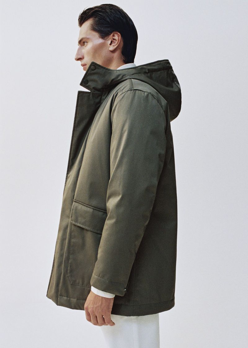 Man in an olive green hooded parka coat standing in a neutral studio. The parka is mid-length with large pockets.