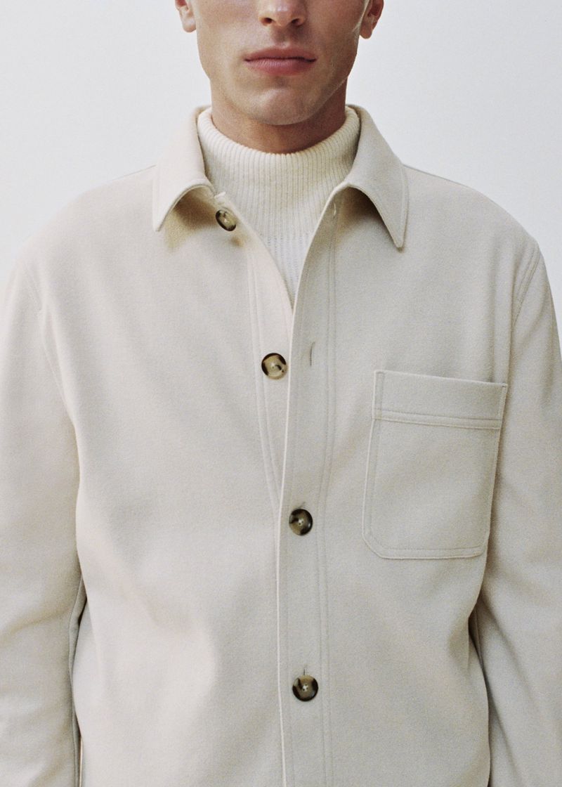 Close-up of a fair-skinned adult male wearing a cream-colored button-down shirt jacket and a cream turtleneck sweater against a white backdrop.