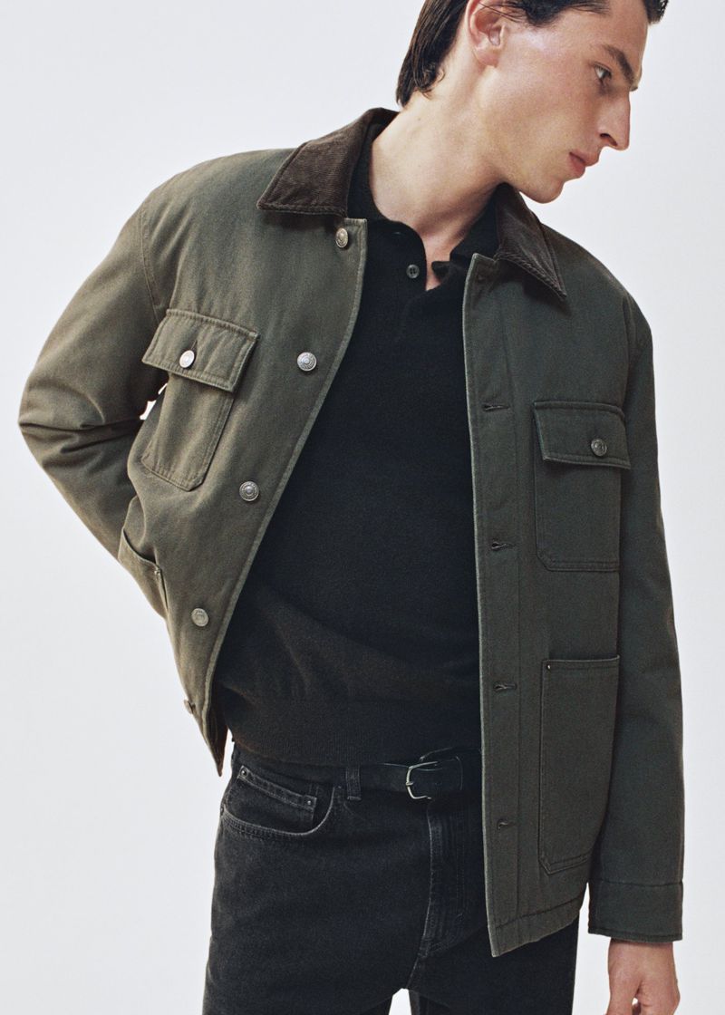 Man poses in olive-green collared workwear jacket with pockets, black polo shirt, jeans and belt against a plain background.