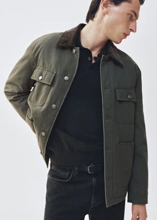 Man poses in olive-green collared workwear jacket with pockets, black polo shirt, jeans and belt against a plain background.