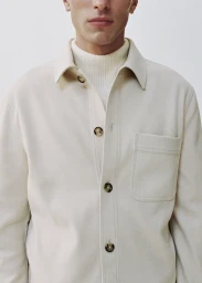 Close-up of a man wearing a cream overshirt and ribbed turtleneck sweater against a neutral background