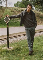 Model leaning against a post beside a rural pathway wearing a grey zip jacket and trousers for Mango Man November 2025.