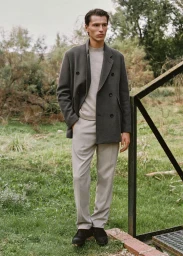 Clément Laguardia wearing a charcoal double-breasted coat and light trousers standing on grass near a black metal railing.