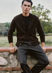 Model Clément Laguardia seated on a low stone wall wearing a dark brown knit cardigan and charcoal trousers for Mango Man.