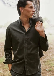 A man wearing a dark button-down shirt outdoors, holding a jacket over his shoulder against a tree and forest background.