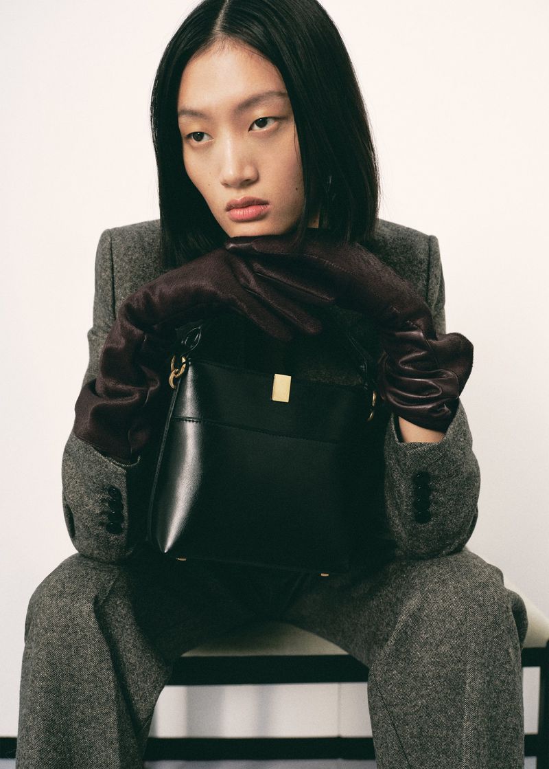 Asian fashion model poses with a black leather purse, wearing gloves and a grey tailored suit, indoors against a plain white wall.