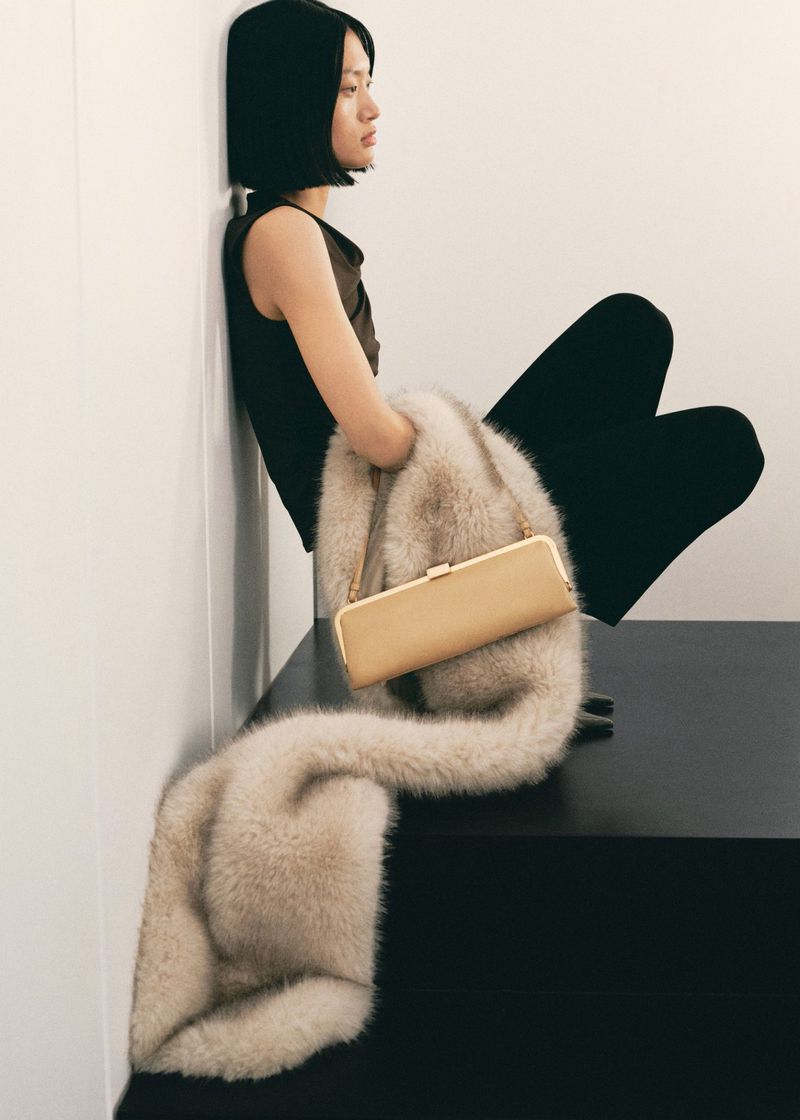 Elegant woman in a dark top and pants is sitting next to a faux fur stole draped over a black bench. A beige handbag is on top.