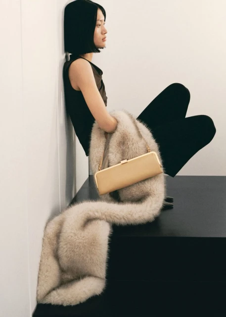 Elegant woman in a dark top and pants is sitting next to a faux fur stole draped over a black bench. A beige handbag is on top.