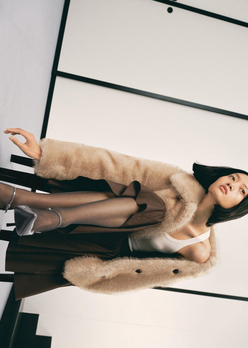 Fashion editorial shot of an Asian woman lying on a black metal bench wearing a light brown coat, brown skirt, and silver heels.