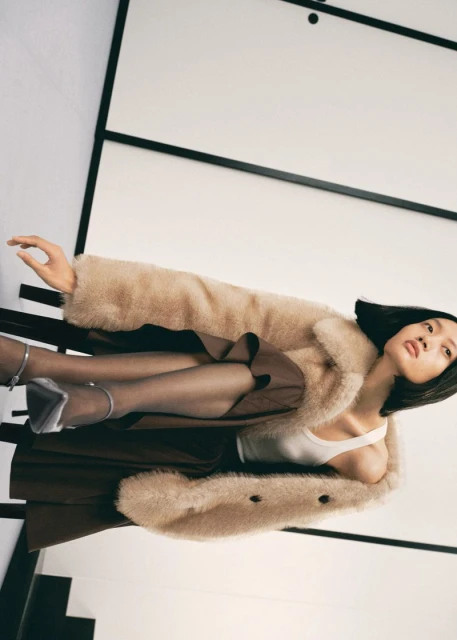 Fashion editorial shot of an Asian woman lying on a black metal bench wearing a light brown coat, brown skirt, and silver heels.