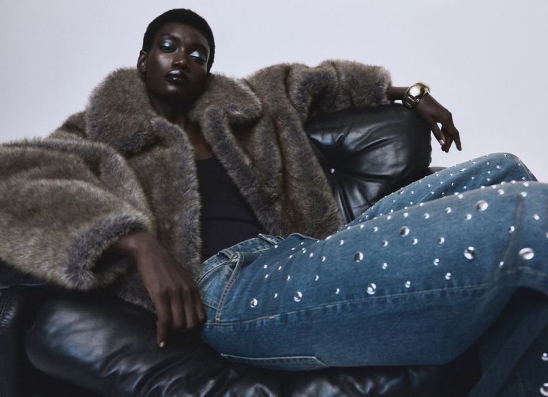 A Black woman poses while reclined in a leather armchair, wearing a faux fur coat and jeans with metal ring accents.