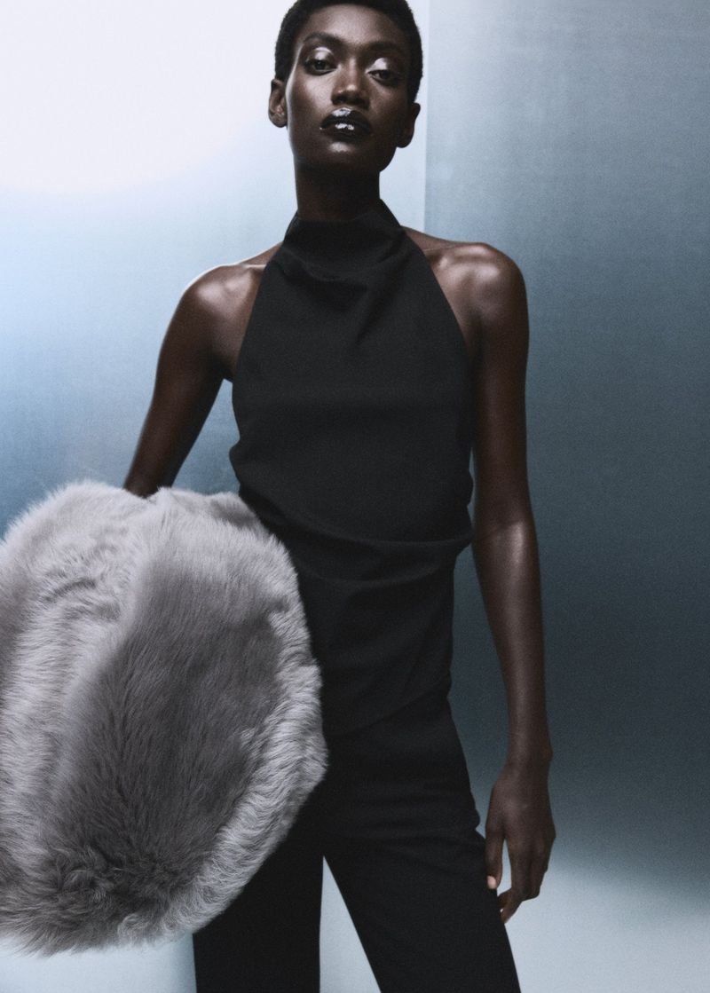 Fashion portrait of a Black woman in black top and pants, holding a grey fur muff against a gradient blue-grey background in studio light.