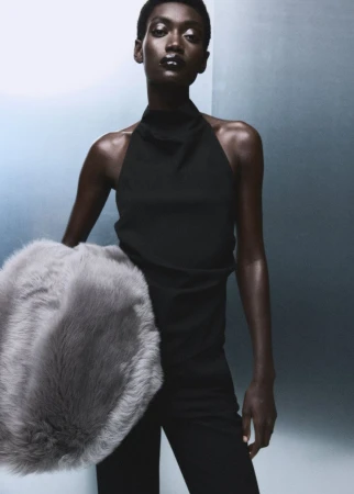 Fashion portrait of a Black woman in black top and pants, holding a grey fur muff against a gradient blue-grey background in studio light.