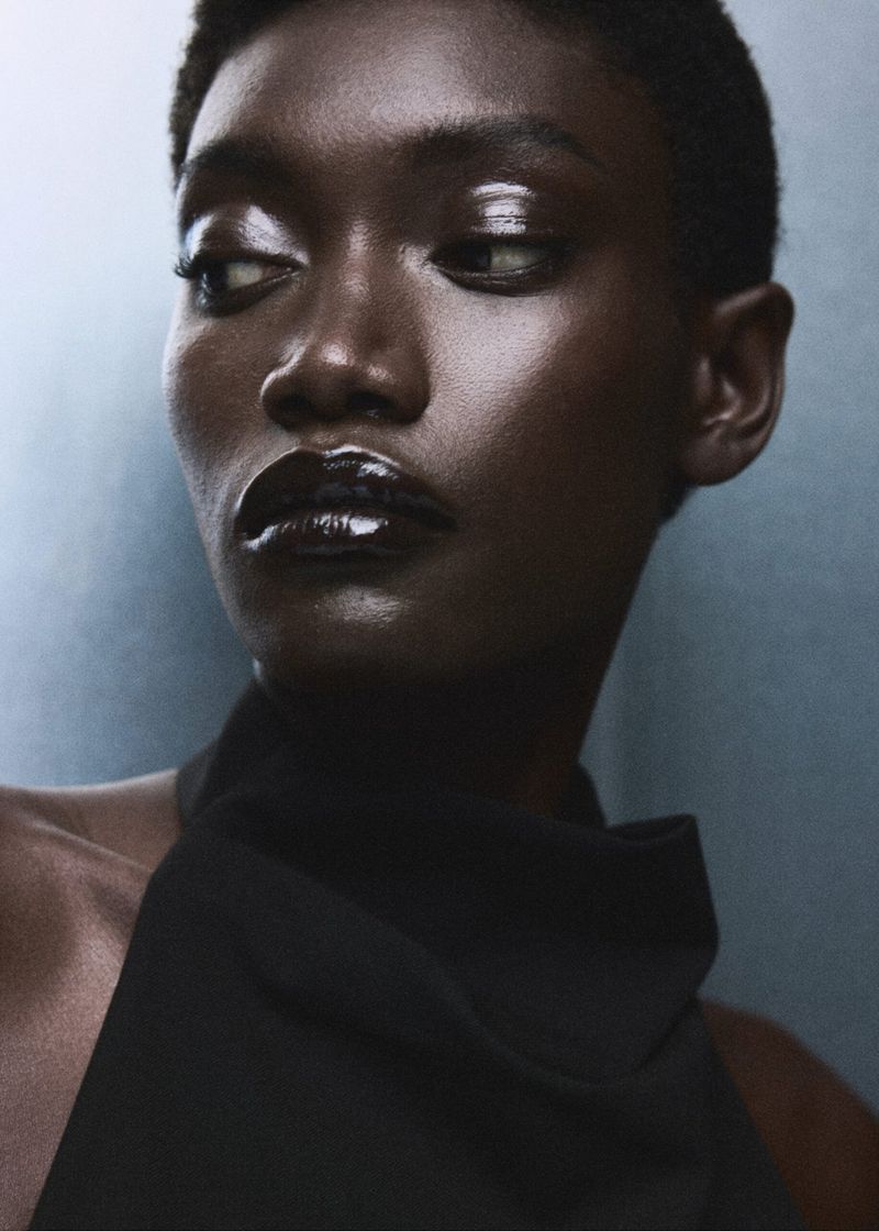 Portrait of a woman with dark skin and short hair wearing black top. The image features glossy makeup with highlights and dark lipstick.