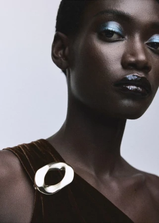 Close-up of a woman with dark skin, wearing a brown, one-shoulder top with silver ring detail. She has light blue eye shadow.