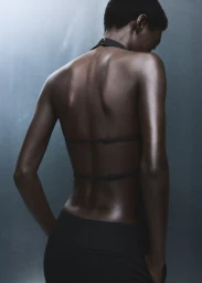 Model Fatou Kebbeh seen from behind wearing a black backless halter top with thin horizontal straps against a cool-toned studio backdrop.
