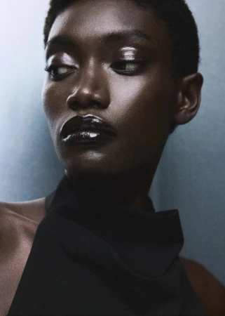 Model Fatou Kebbeh wearing a black high-neck top with glossy lips and dewy skin against a soft cool-toned studio backdrop.