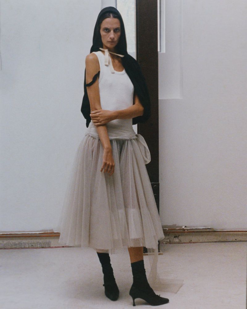 A slender woman with dark hair stands in a studio wearing a white tank top, tulle skirt, black socks, and heeled shoes.