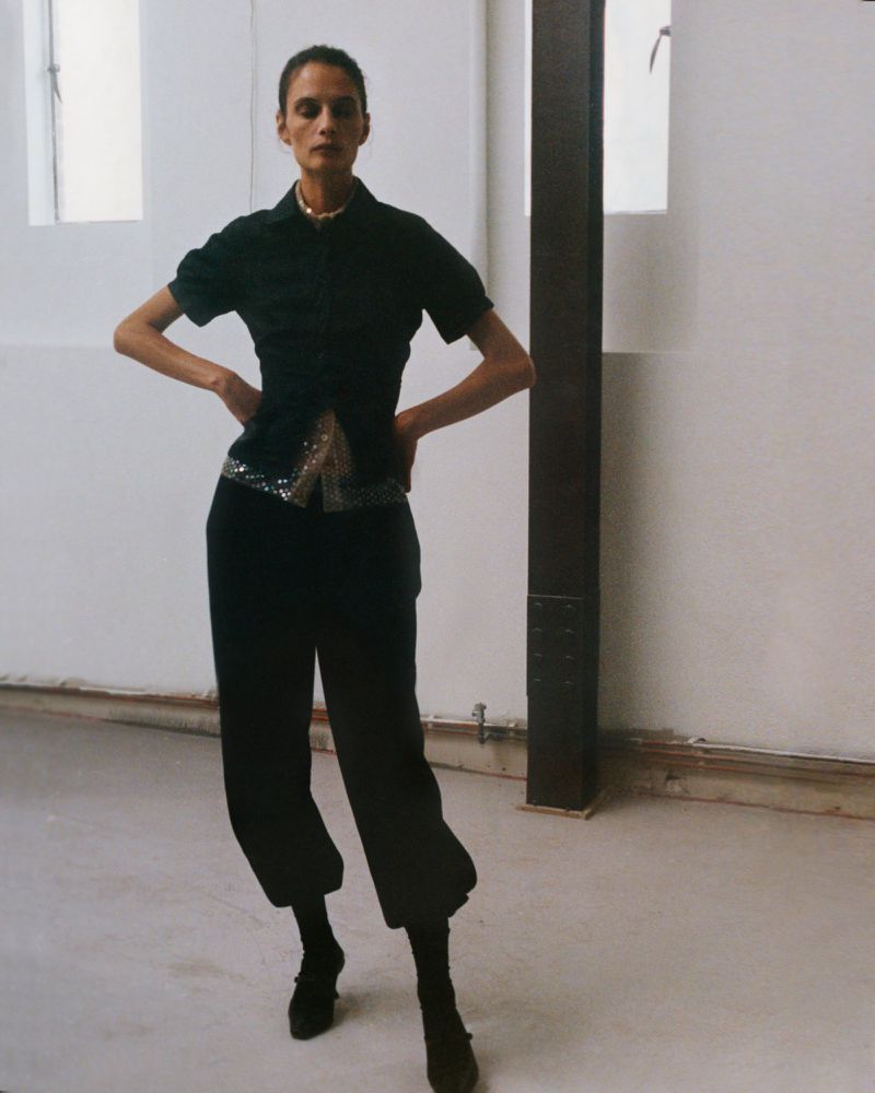 An adult female model poses in a black top and pants with a silver belt in an indoor studio with neutral colors and natural lighting.