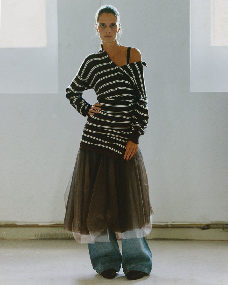 Fashionable woman poses in a black and white striped sweater, tulle skirt, and jeans against a white wall, looking confidently at the camera.