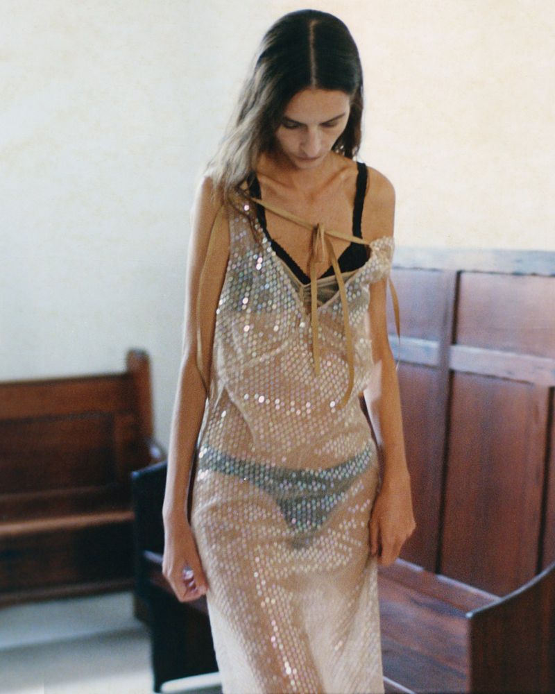 Brunette woman in a sheer sequin dress stands indoors with pews in the background. The image has a slightly desaturated, vintage tone.