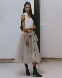 Model Suzie Bird standing in a white tank top, sheer beige tulle midi skirt, black headpiece, and black kitten-heel sock boots.