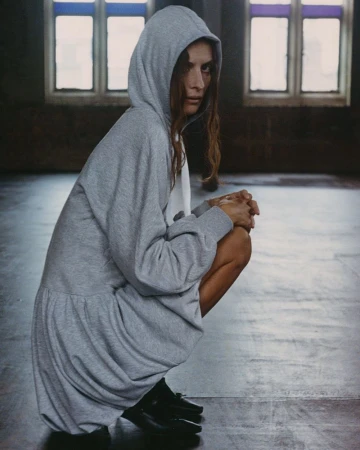 Model Suzie Bird crouching in a grey hooded fleece dress with black shoes for Renaissance Renaissance.