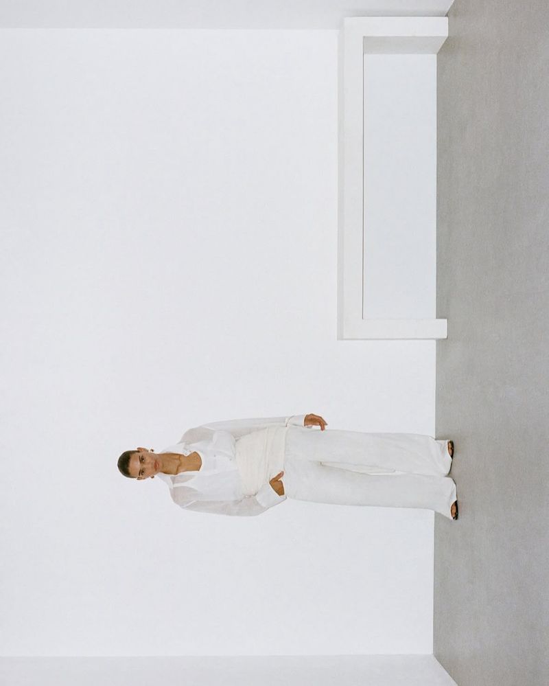 Woman in white linen outfit poses in a white-walled corner with grey floor and minimalist shelf, captured in high-key lighting.