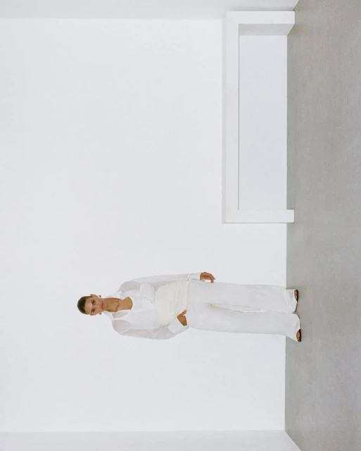 Woman in white linen outfit poses in a white-walled corner with grey floor and minimalist shelf, captured in high-key lighting.