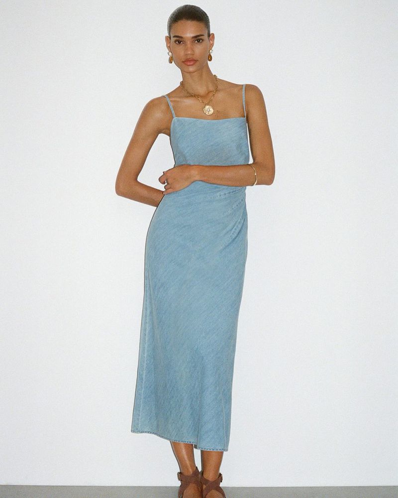 Elegant adult female model in a light blue midi dress, posing in a full shot against a bright white backdrop in a fashion style.