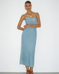 Model Barbara Valente wearing a light blue denim midi dress and gold jewelry against a white studio background.
