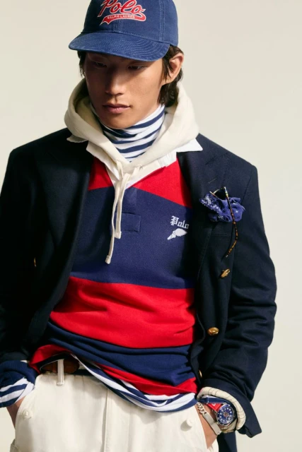 Asian male model wearing a navy blue blazer, hoodie, striped turtleneck, rugby shirt, baseball cap, and white pants in a studio setting.