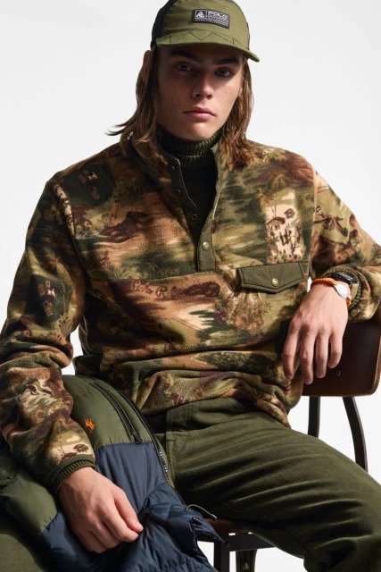 Man in olive green cap and camouflage-pattern fleece pullover poses indoors. Chair and puffer jacket provide additional background.