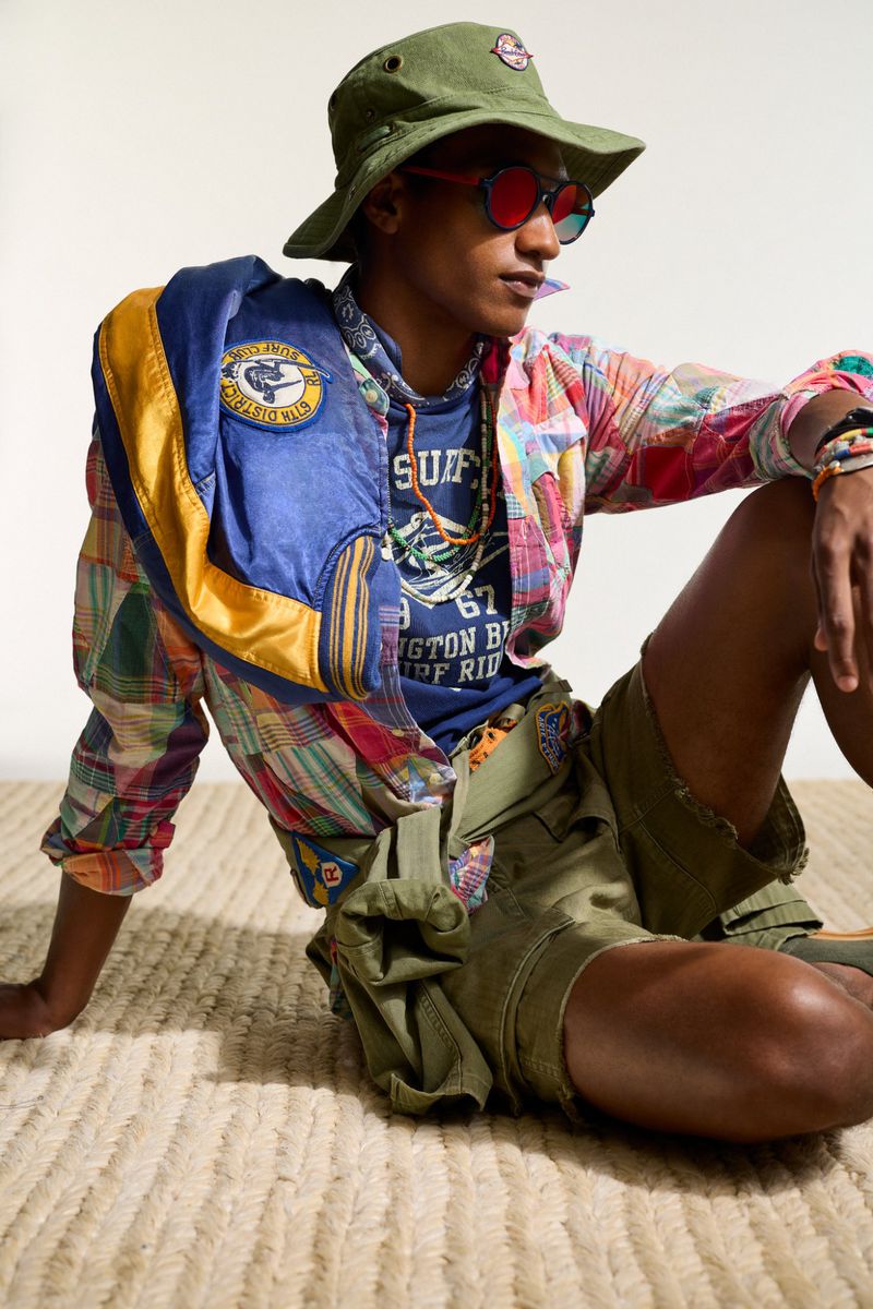 Young adult male in casual wear, including a bucket hat, blue t-shirt, and plaid jacket, is kneeling on a beige, textured rug.