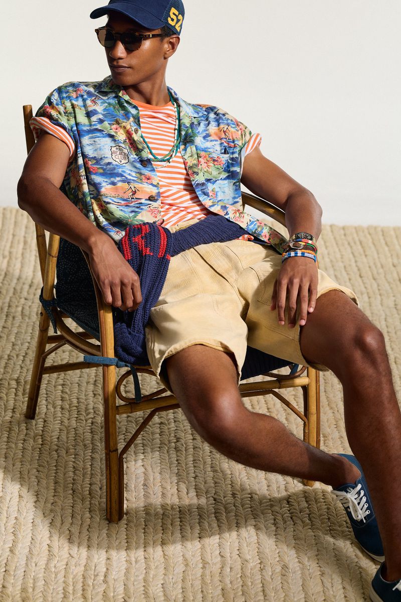 Man in casual summer fashion sits in a bamboo chair wearing a floral shirt, shorts, baseball cap, sunglasses, necklace and bracelets. 